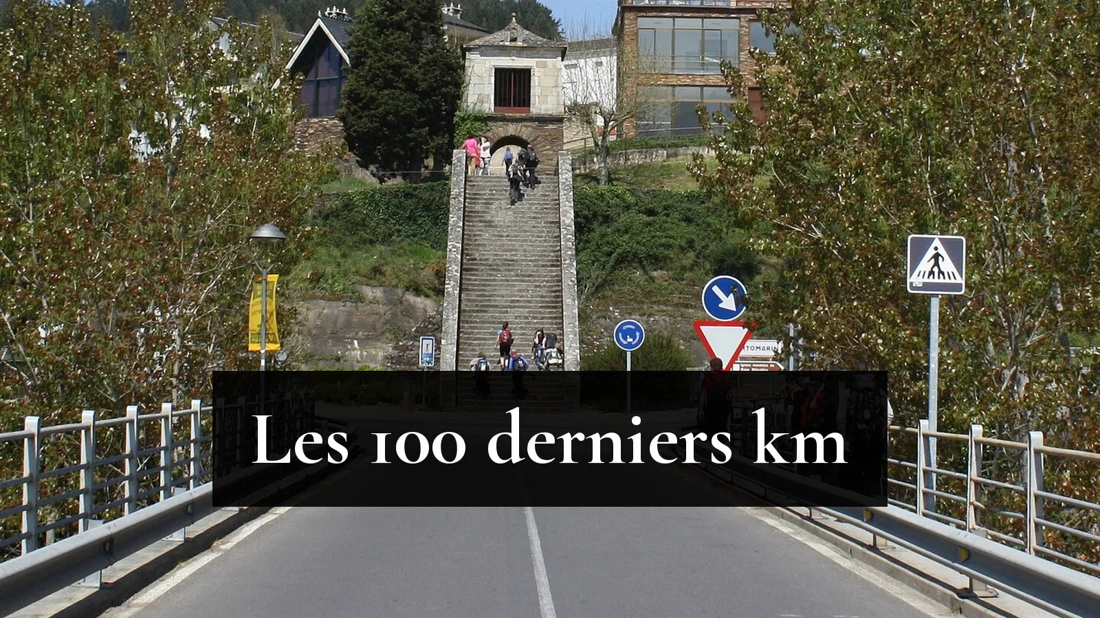 Des pèlerins sac au dos traversent le pont sur le Miño et montent le grand escalier de pierre qui mène à Portomarín, sous un ciel bleu